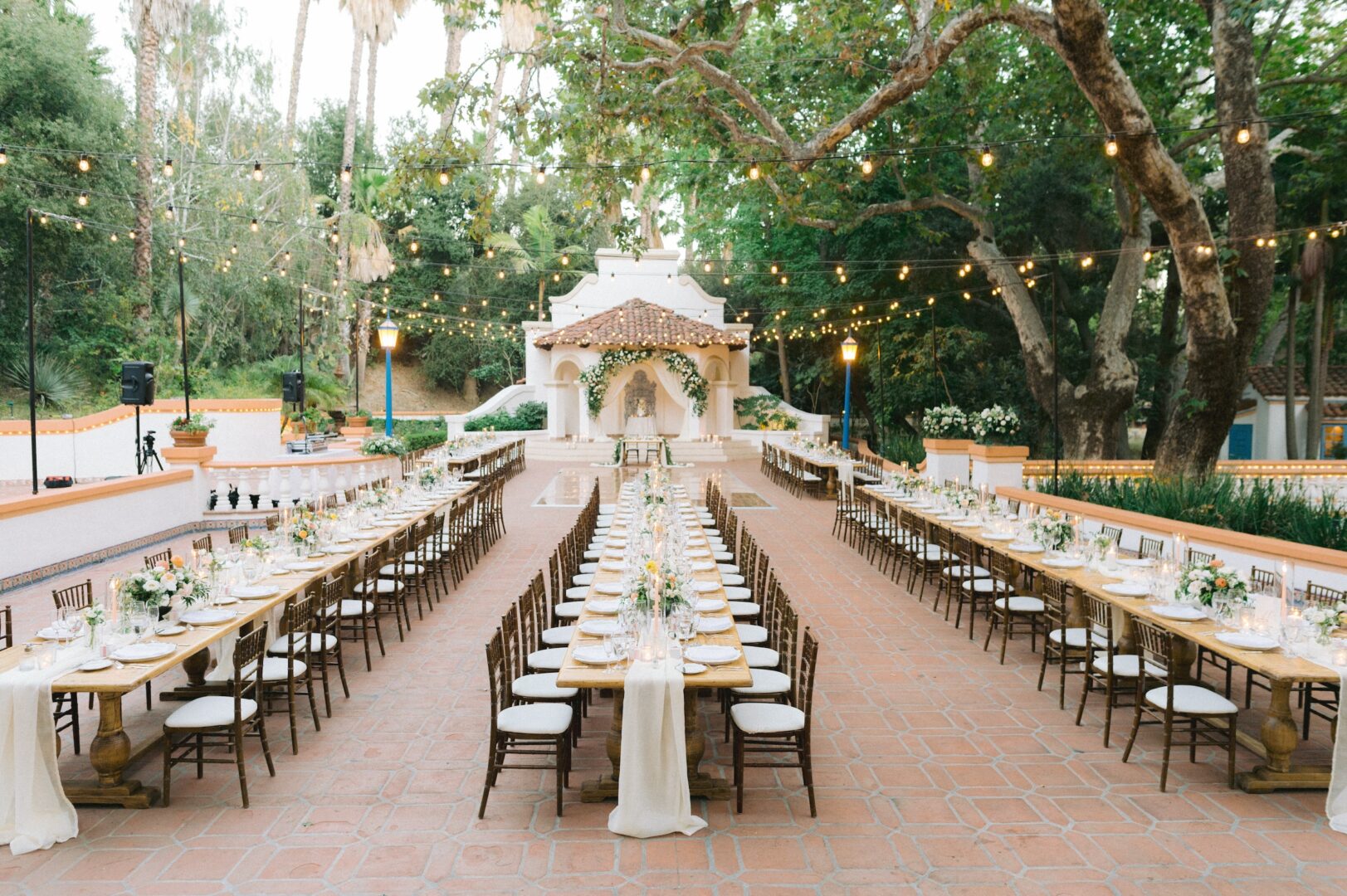 Long banquet tables with elegant place settings are arranged outdoors under string lights, facing a small decorated altar surrounded by greenery and trees, creating a festive wedding atmosphere.