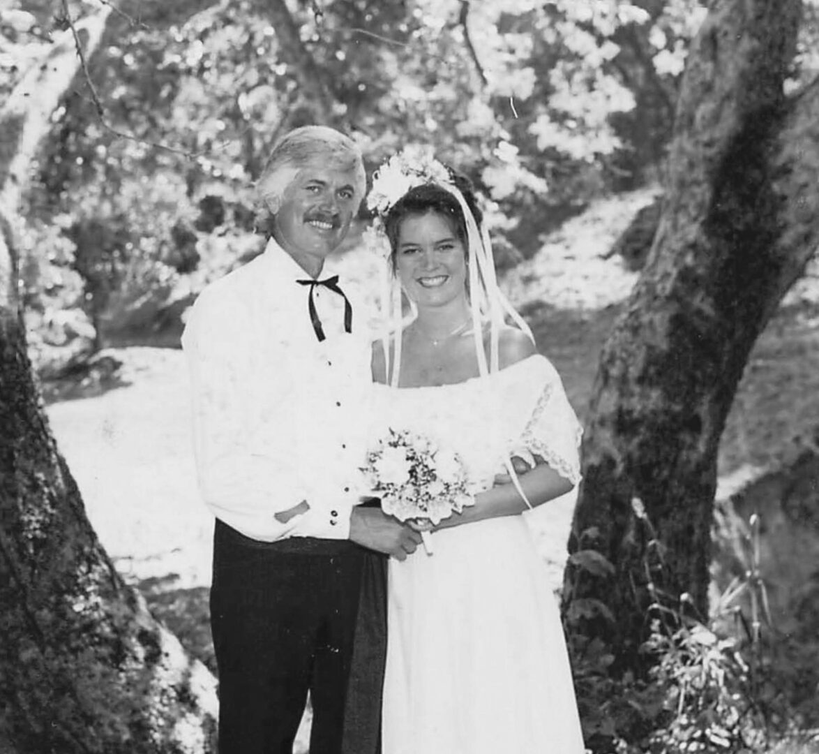 A smiling bride and groom stand together outdoors among trees, with the bride holding a bouquet and the groom wearing a bow tie.