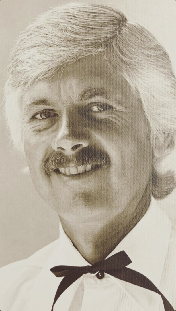 A black-and-white portrait of a smiling man with light hair, a mustache, and a bow tie, dressed in a collared shirt.