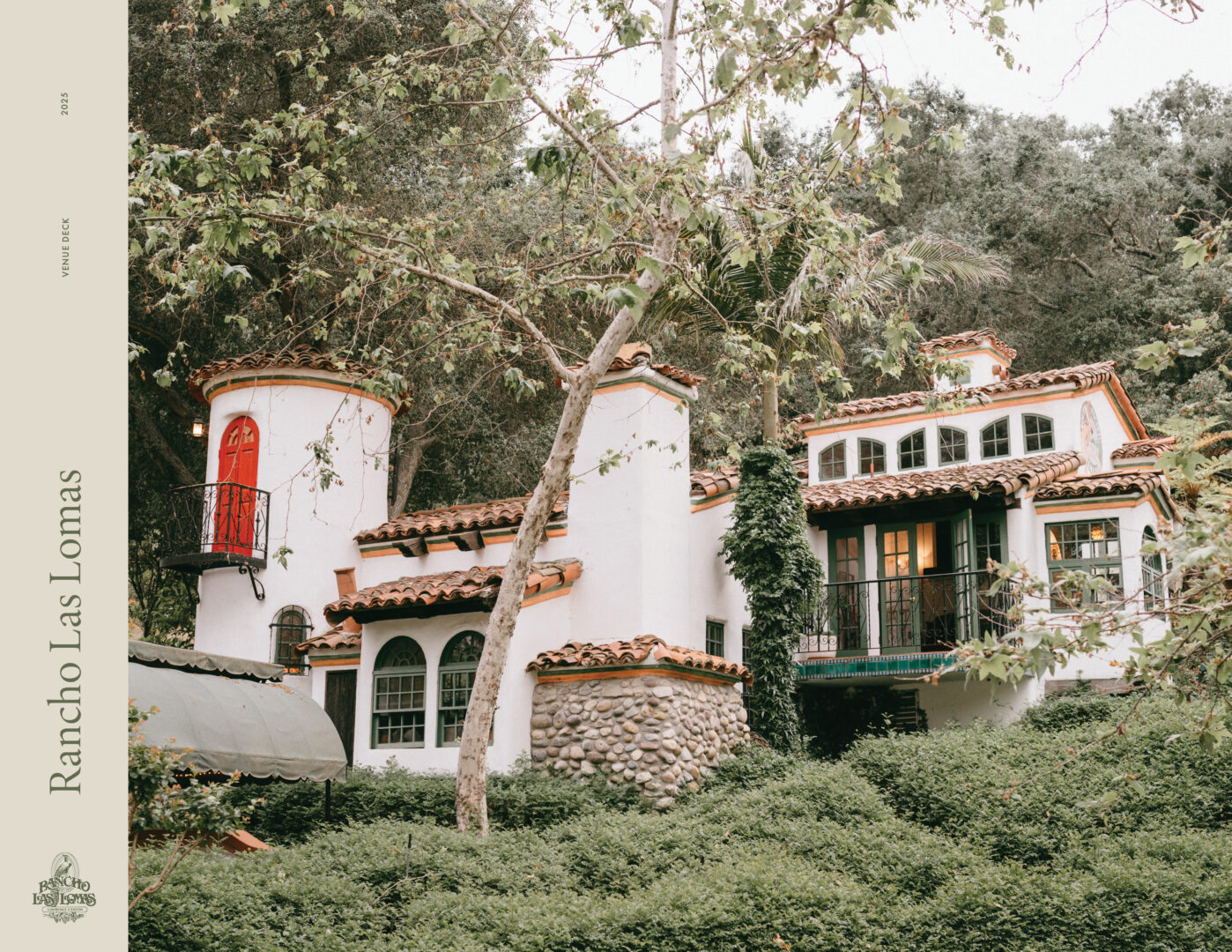 A Spanish-style villa with white walls, terracotta roof tiles, arched windows, and a red door, surrounded by lush greenery and tall trees. Text on the left reads Rancho Las Lomas..