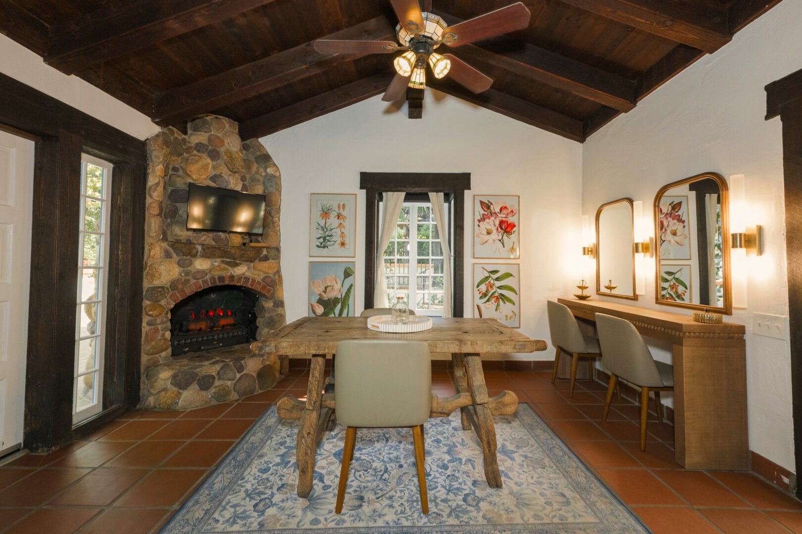 Rustic dining room with wood ceiling, stone fireplace, wooden table, four chairs, floral wall art, vanity with mirrors, blue patterned rug, and ceiling fan. Light streams in from a window with white curtains.
