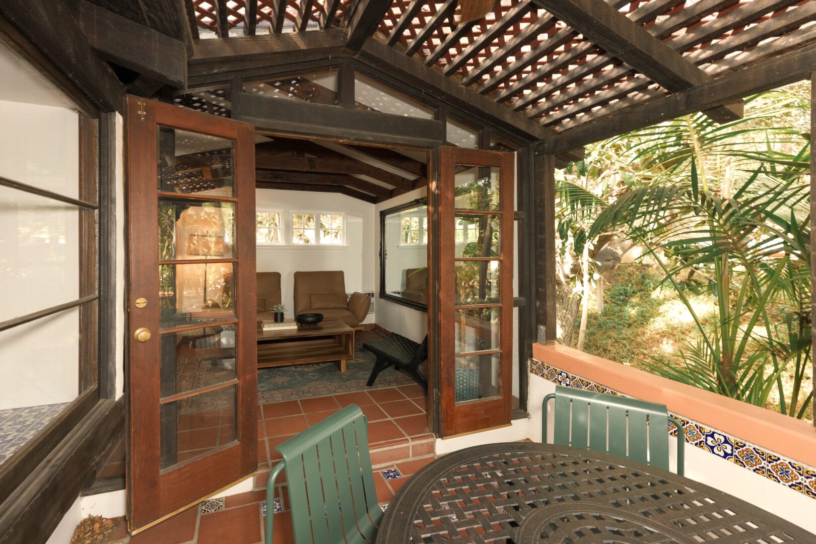 Covered patio with a black metal table and green chairs, open wooden French doors, and a view into a cozy sitting room with lounge chairs and a coffee table. Tropical plants and trees are visible outside.